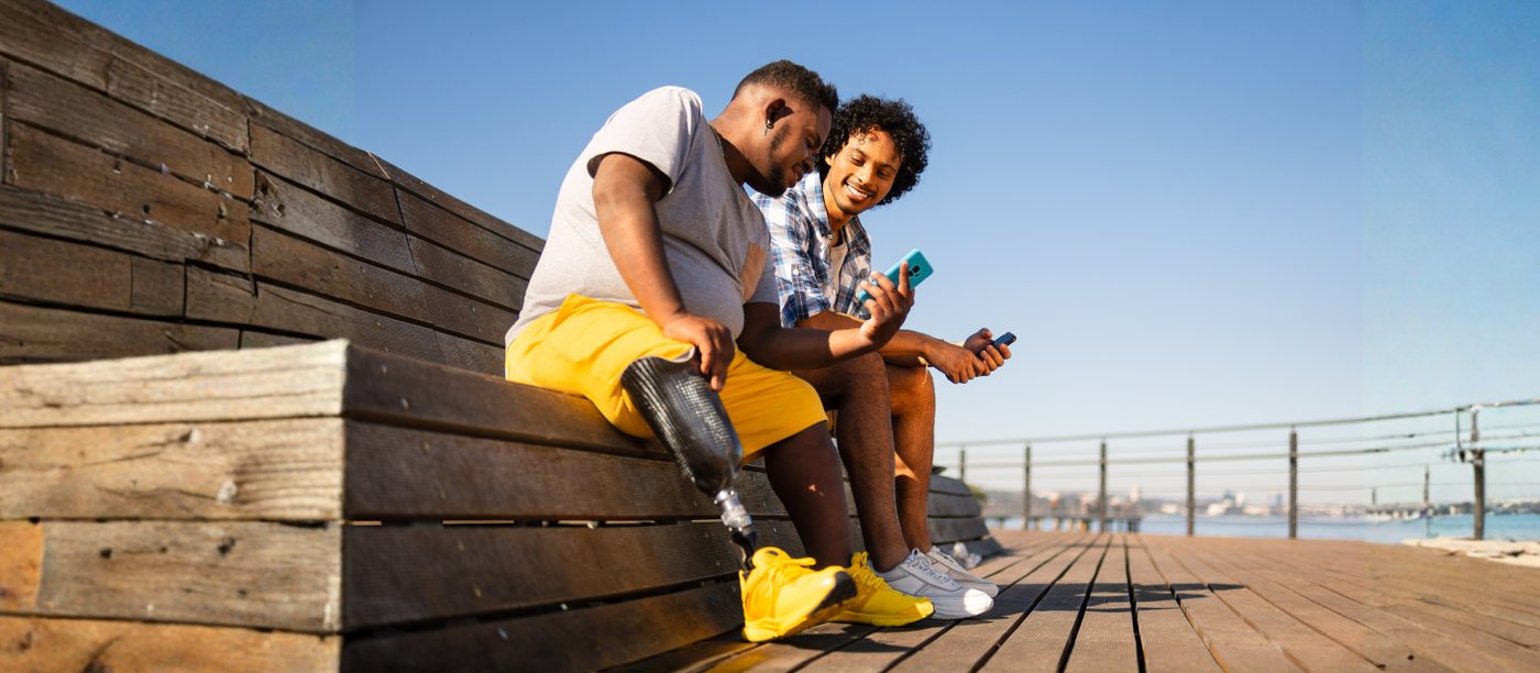 Two people sitting together on a boardwalk in the sun, one with a prosthetic leg, looking at a phone