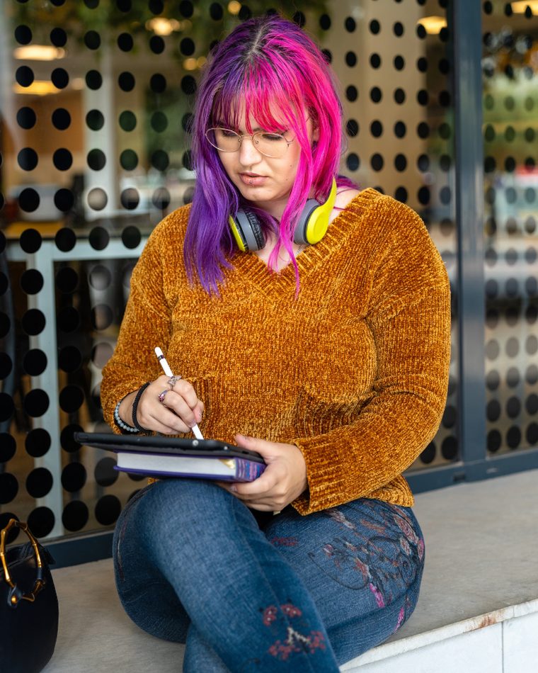 A person with brightly coloured hair sitting and writing, headphones resting around their neck