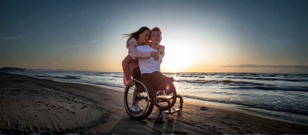 A couple sitting close together at sunset, one using a wheelchair